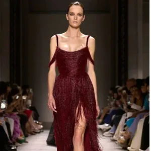 Modelo desfilando con un vestido burdeos de la colección 2024 de Elie Saab, con detalles de flecos y corte con abertura, reflejando un diseño de alta costura sofisticado.